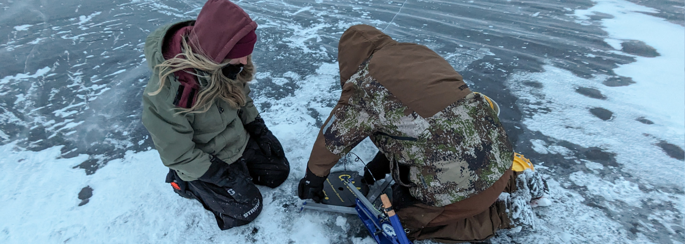 two people dressed for cold weather drilling a hole in the ice