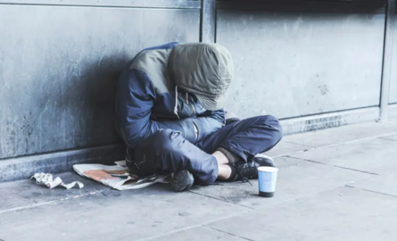 a man with a hood pulled over his face huddles against a wall